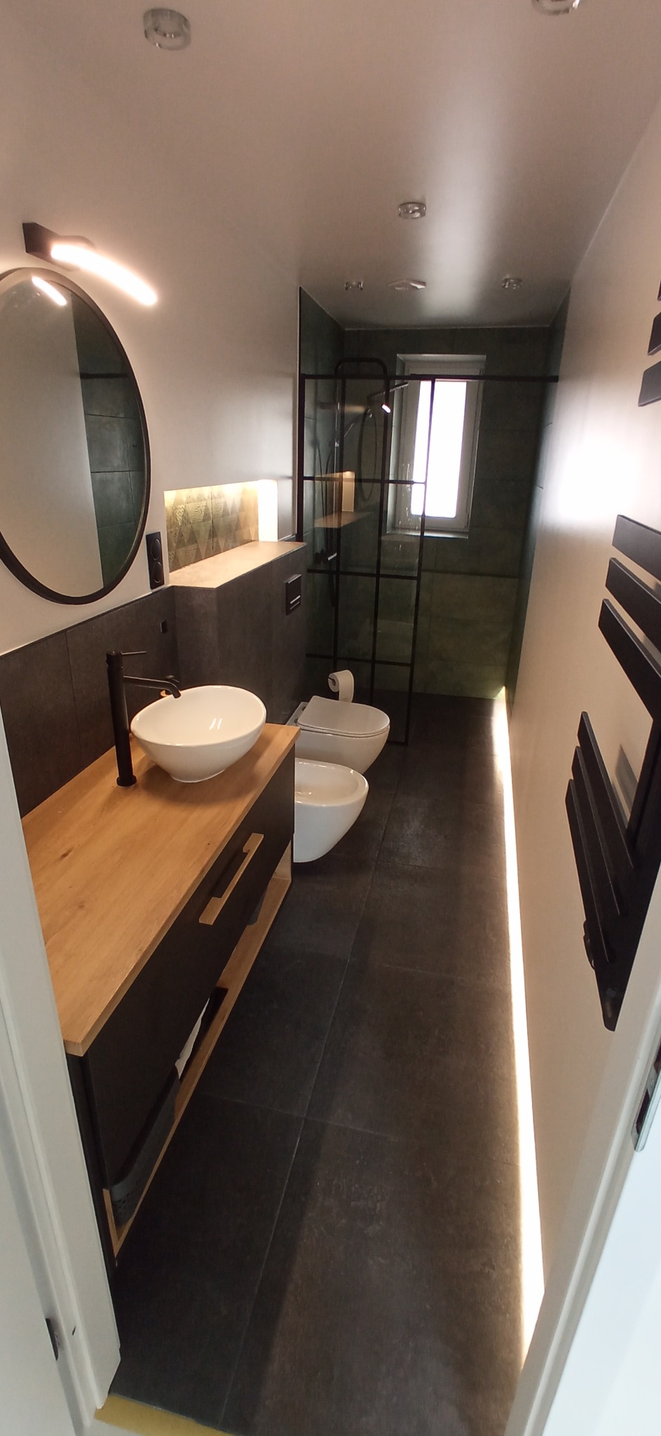 Nowoczesna łazienka z drewnianym blatem pod umywalką nablatową, czarną armaturą, toaletą i bidetem w tle, podświetlona listwą LED wzdłuż ściany.