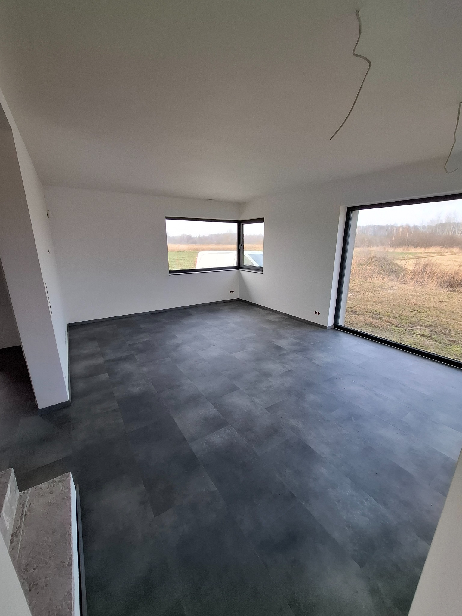 Nowoczesne wnętrze z szarą podłogą panelową i dużymi oknami z widokiem na krajobraz, widoczne kable wystające z sufitu.