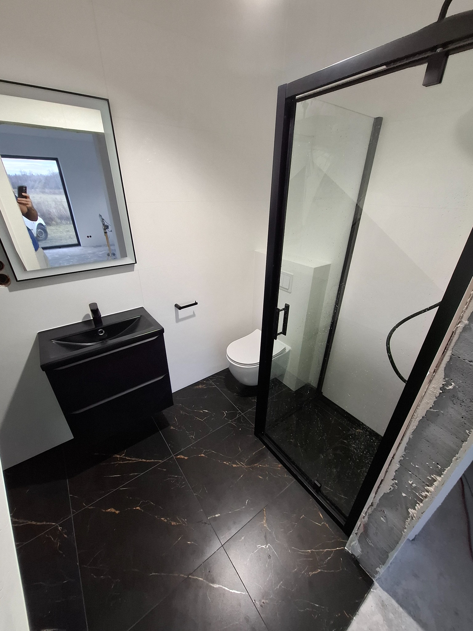 Nowoczesna łazienka z czarną armaturą i płytkami imitującymi marmur, kabina prysznicowa z czarnymi ramami, biała toaleta podwieszana, odbicie fotografa w lustrze.