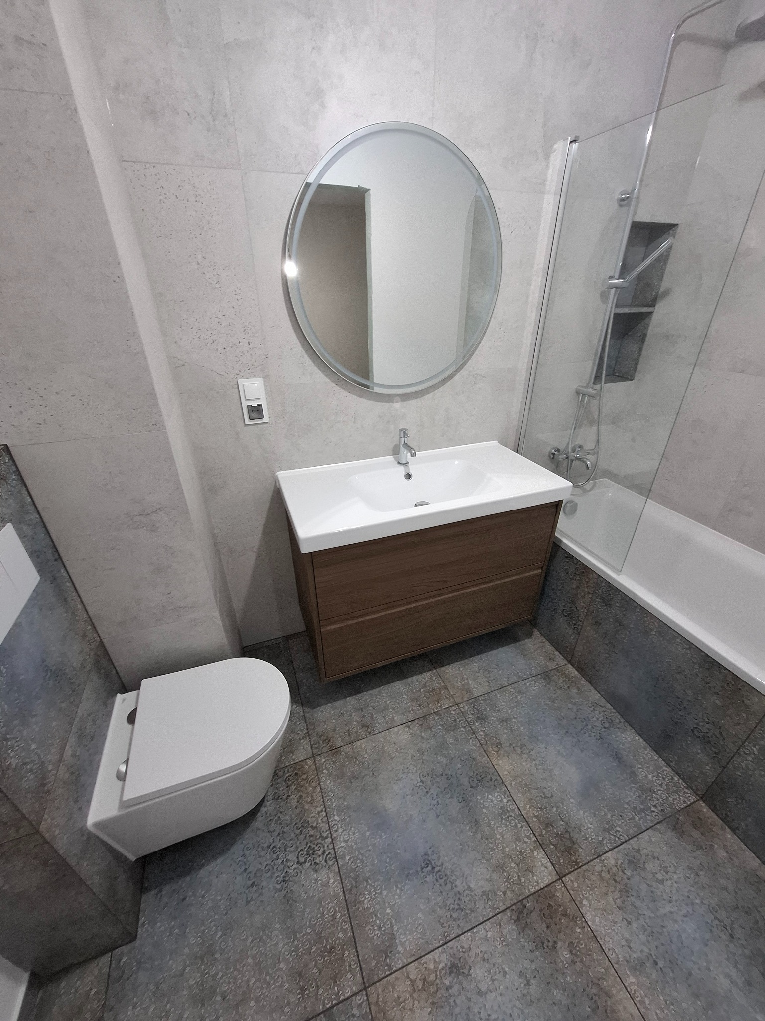 Nowoczesna łazienka z owalnym lustrem, umywalką z drewnianą szafką, toaletą i kabiną prysznicową. Szare płytki na ścianach i podłodze z delikatnym wzorem.