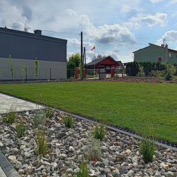 Blossom Garden - Widok na świeżo założony trawnik z obrzeżem z kamieni i roślin, w tle domy i maszt z polską flagą.