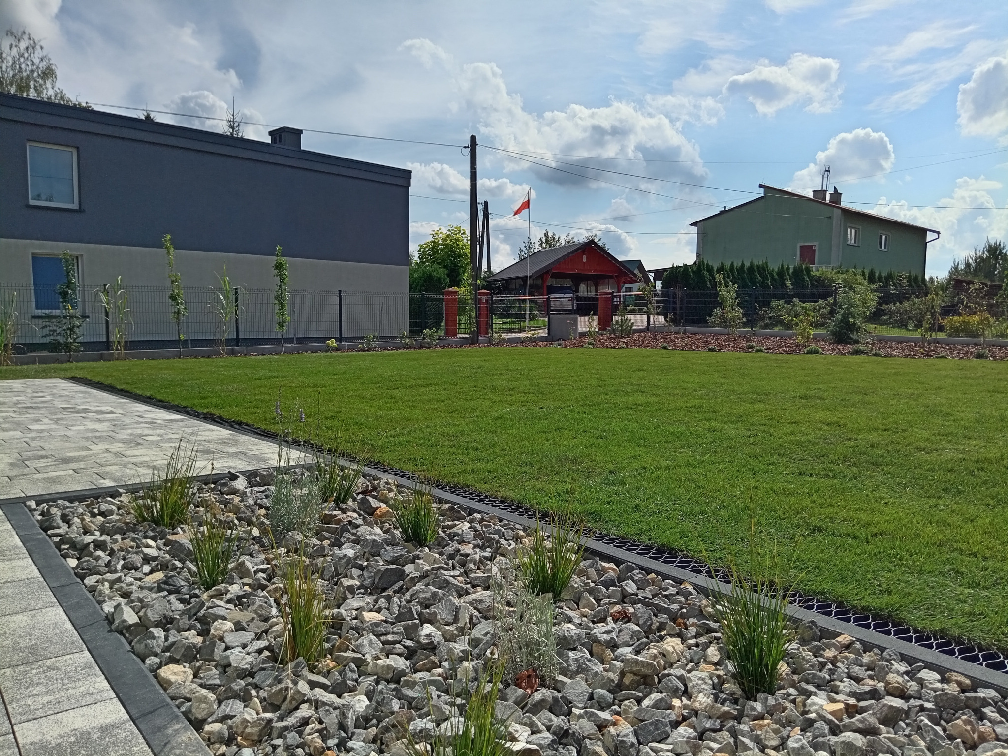 Widok na świeżo założony trawnik z obrzeżem z kamieni i roślin, w tle domy i maszt z polską flagą.