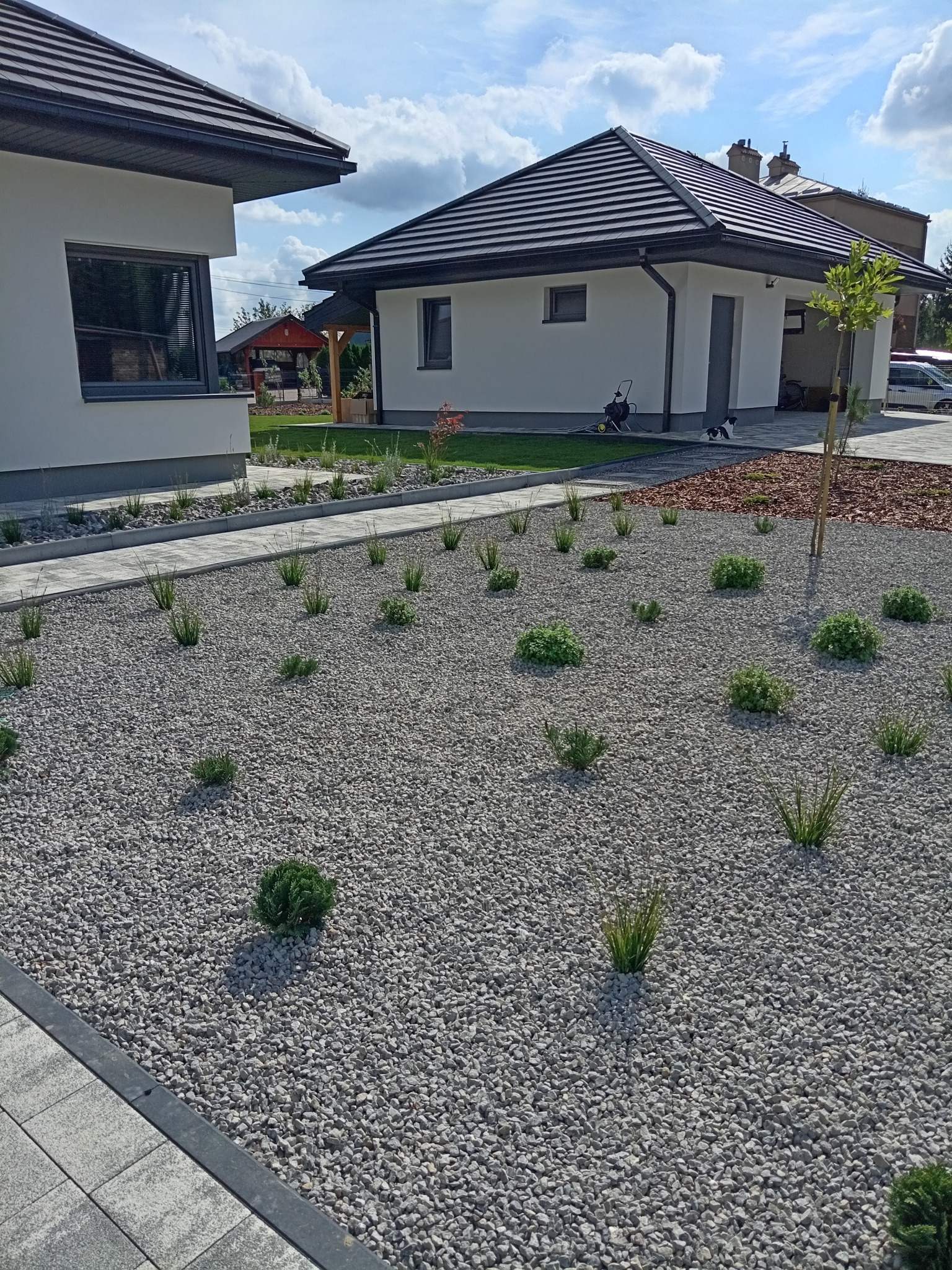 Nowoczesny ogród przydomowy z białym żwirem, niskimi roślinami ozdobnymi i ścieżką z kostki brukowej, w tle dwa białe domy z ciemnymi dachami i elementami małej architektury ogrodowej.