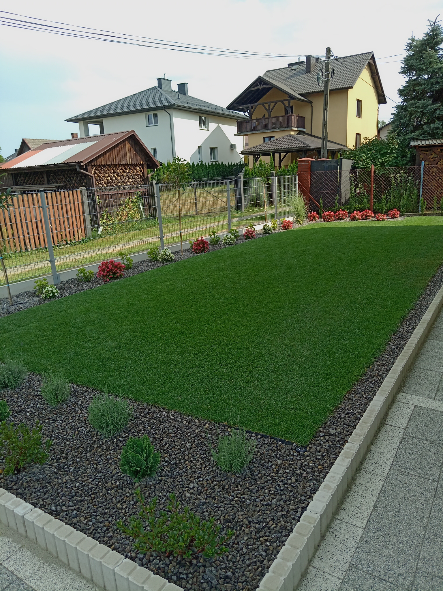 Zadbany trawnik z rolki, obrzeża z kamieni i betonowych krawężników, rabaty z kwiatami wzdłuż ogrodzenia z siatki, domy w tle.