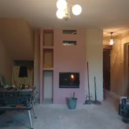 Wnętrze w trakcie remontu: zabudowa kominka z płyt kartonowo-gipsowych z wkładem kominkowym, widoczne narzędzia, wiadro i prowizoryczne półki.