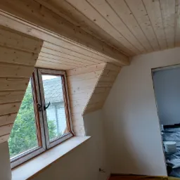 Wykończone drewnem poddasze z oknem, widoczne deski na suficie i ścianach wokół okna, podłoga zabezpieczona folią, przygotowanie do malowania ścian.