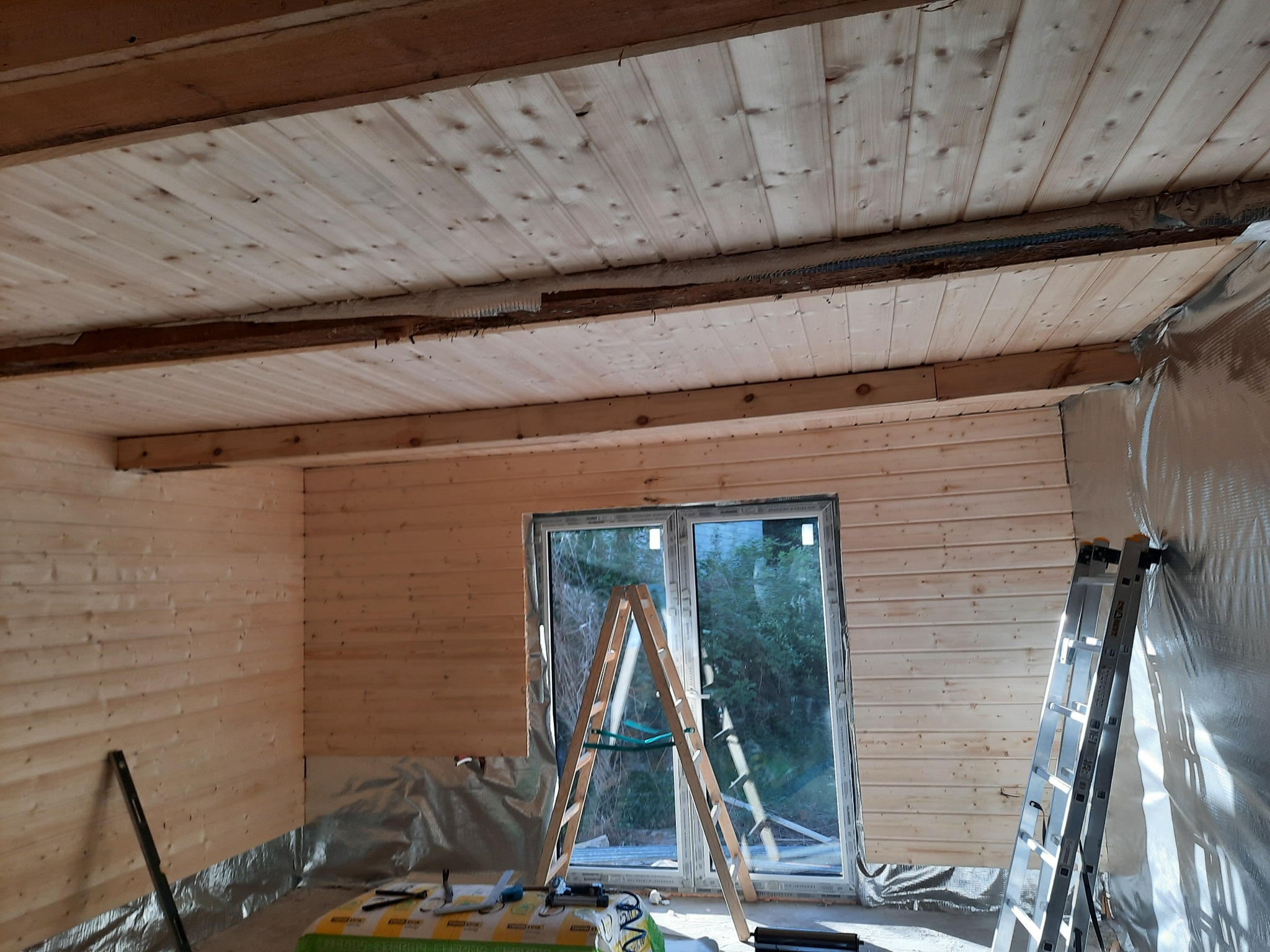 Wnętrze poddasza w trakcie wykańczania boazerią, widoczne fragmenty izolacji, okno, drabina i narzędzia na podłodze zabezpieczonej folią.
