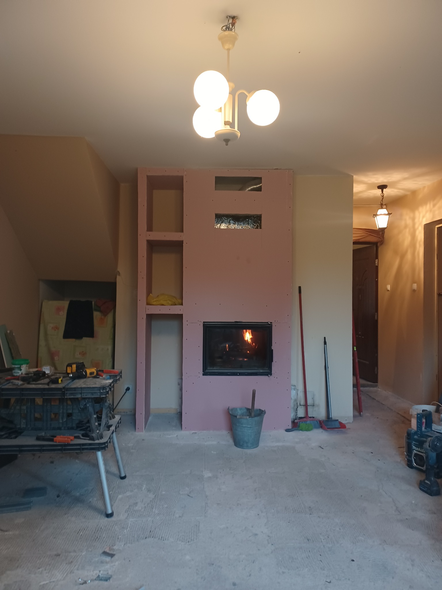 Wnętrze w trakcie remontu: zabudowa kominka z płyt kartonowo-gipsowych z wkładem kominkowym, widoczne narzędzia, wiadro i prowizoryczne półki.
