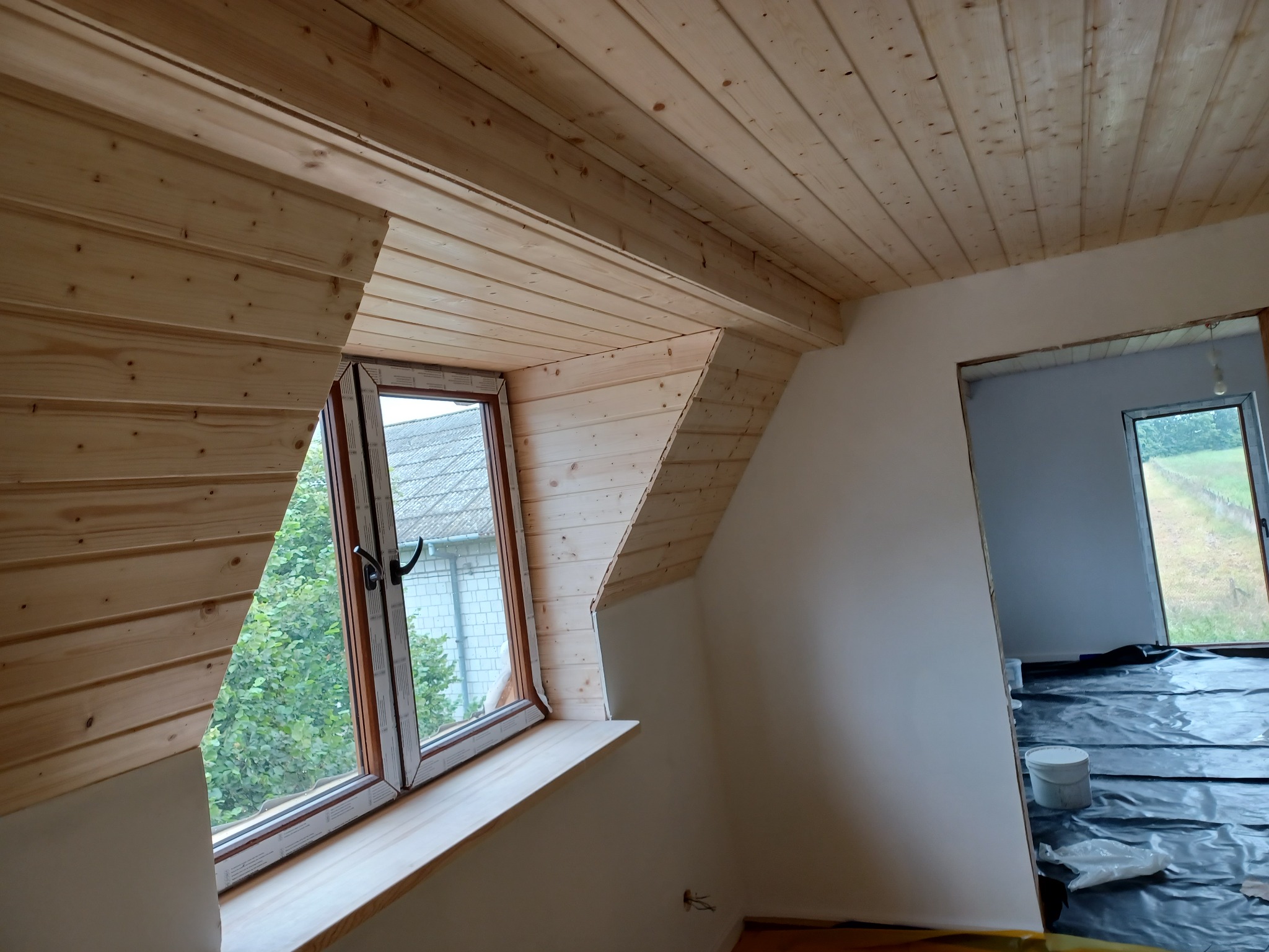Wykończone drewnem poddasze z oknem, widoczne deski na suficie i ścianach wokół okna, podłoga zabezpieczona folią, przygotowanie do malowania ścian.