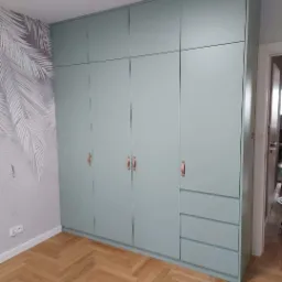 Duża, pastelowo-zielona szafa w zabudowie z drewnianymi uchwytami na tle ściany z motywem liści palmowych i parkietu ułożonego w jodełkę.