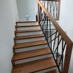 Nowoczesne schody z drewnianymi stopniami i czarną balustradą o geometrycznym wzorze, widoczny pojemnik z farbą na górnym stopniu.