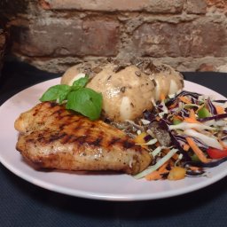 PHU Anna Jarmuż - Grillowana pierś kurczaka, trzy ziemniaki w sosie grzybowym i kolorowa surówka na różowym talerzu, bazylia jako dekoracja, na tle ściany z cegły.