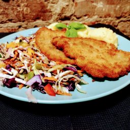 PHU Anna Jarmuż - Dwa panierowane kotlety na turkusowym talerzu, podane z puree ziemniaczanym i surówką warzywną; całość udekorowana listkami bazylii, na tle ceglanej ściany.