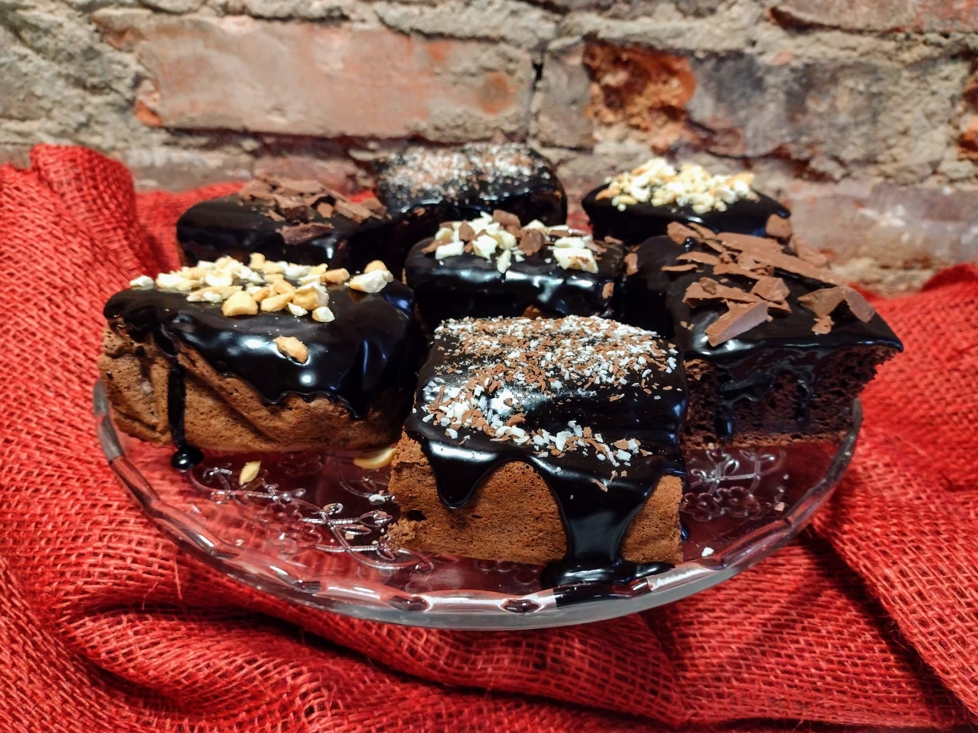 Siedem kawałków brownie z polewą czekoladową i różnymi posypkami (orzechy, wiórki kokosowe, czekolada) na szklanym talerzu z dekoracyjnym brzegiem, na tle czerwonej tkaniny i ceglanej ściany.