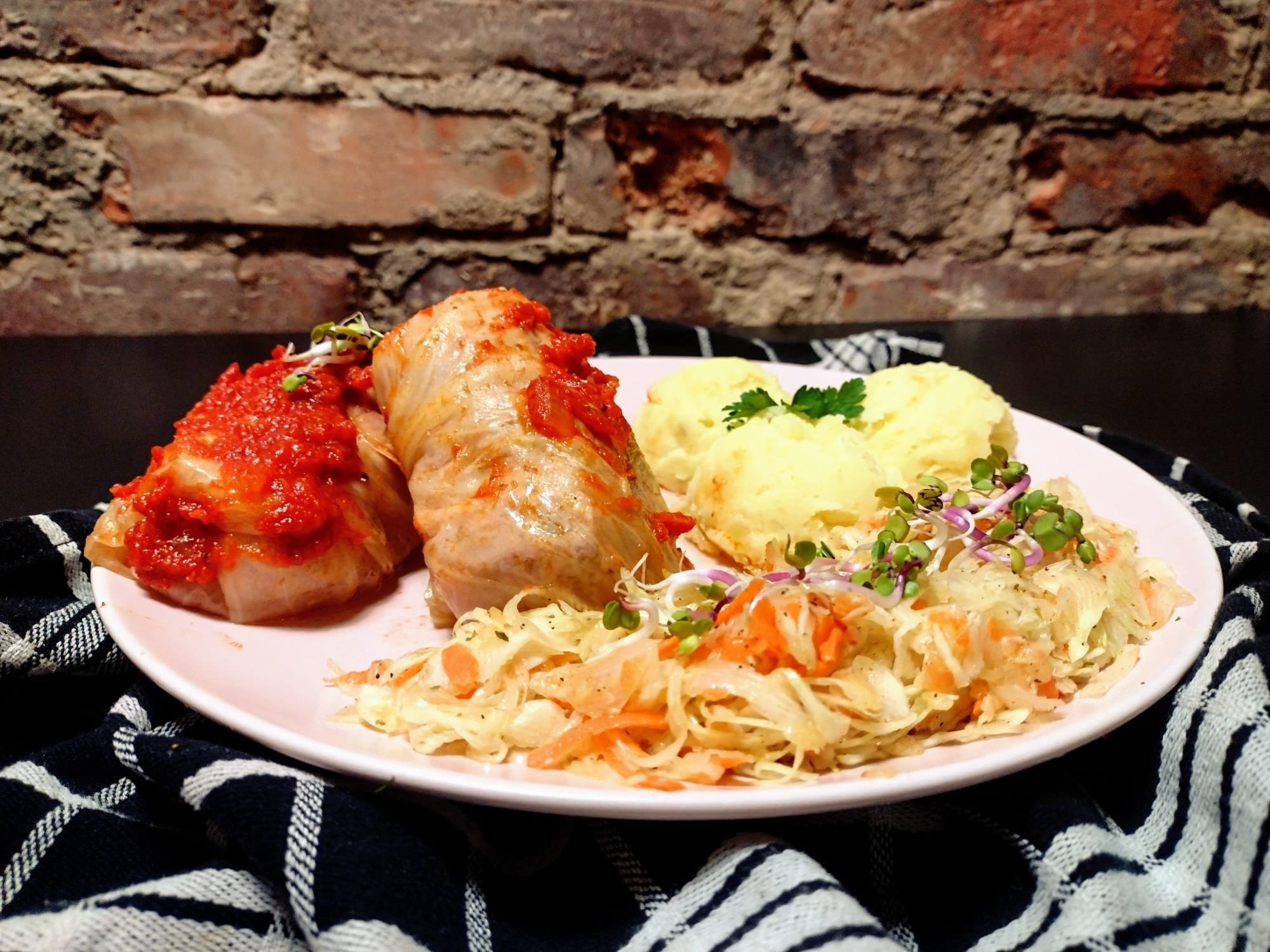 Trzy gołąbki polane sosem pomidorowym, puree ziemniaczane i surówka z kapusty na różowym talerzu, na tle ceglanej ściany i czarno-białej ściereczki.
