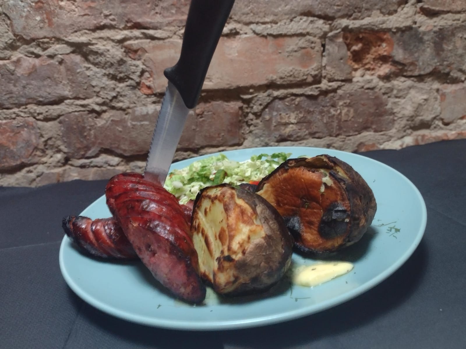 Grillowana kiełbasa, pieczone ziemniaki z sosem i sałatka na niebieskim talerzu, nóż wbity w kiełbasę, ceglana ściana w tle.
