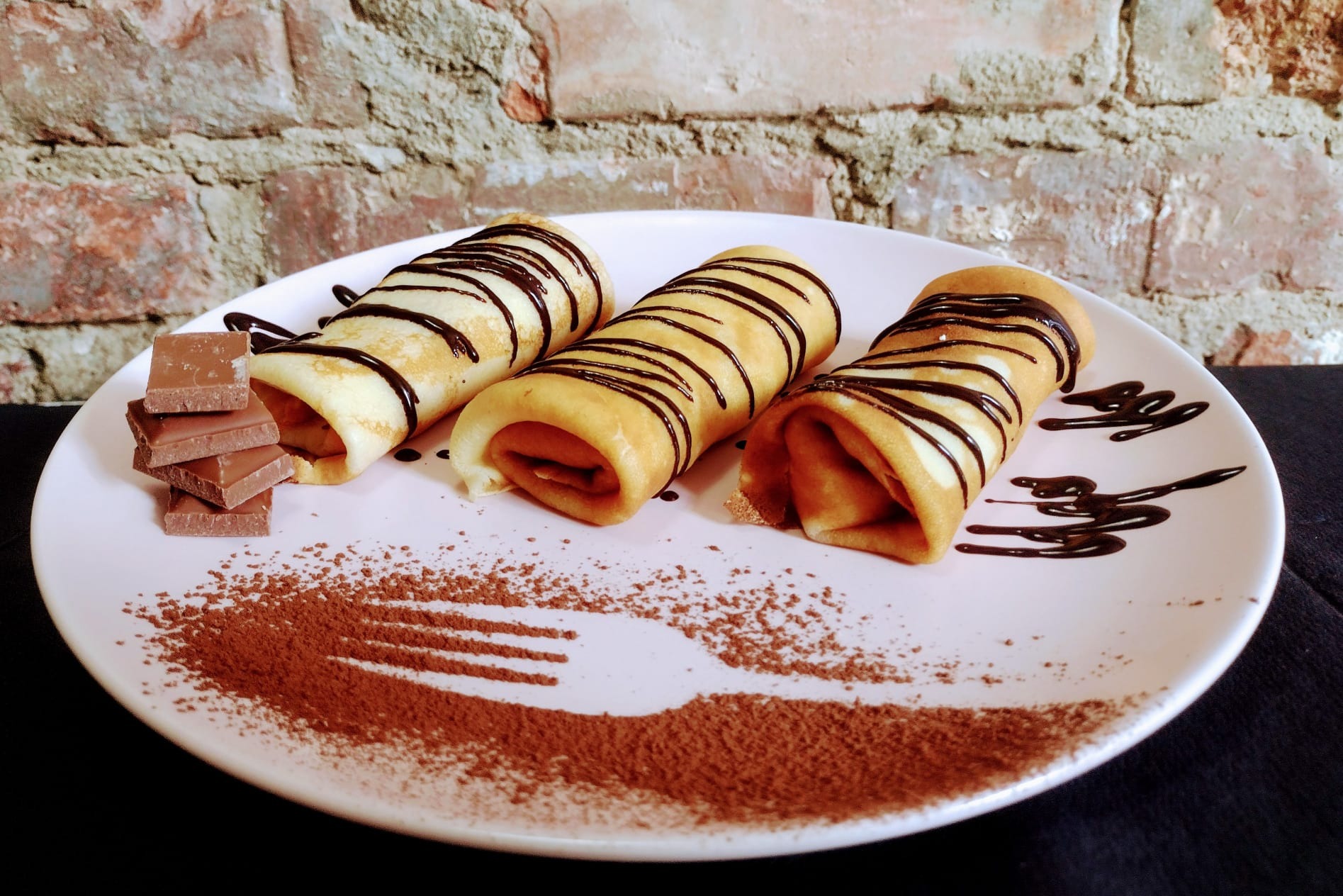 Trzy naleśniki zawinięte z nadzieniem, polane czekoladą, ułożone na talerzu z dekoracją z kakao w kształcie widelca i kostkami czekolady, na tle ceglanej ściany.