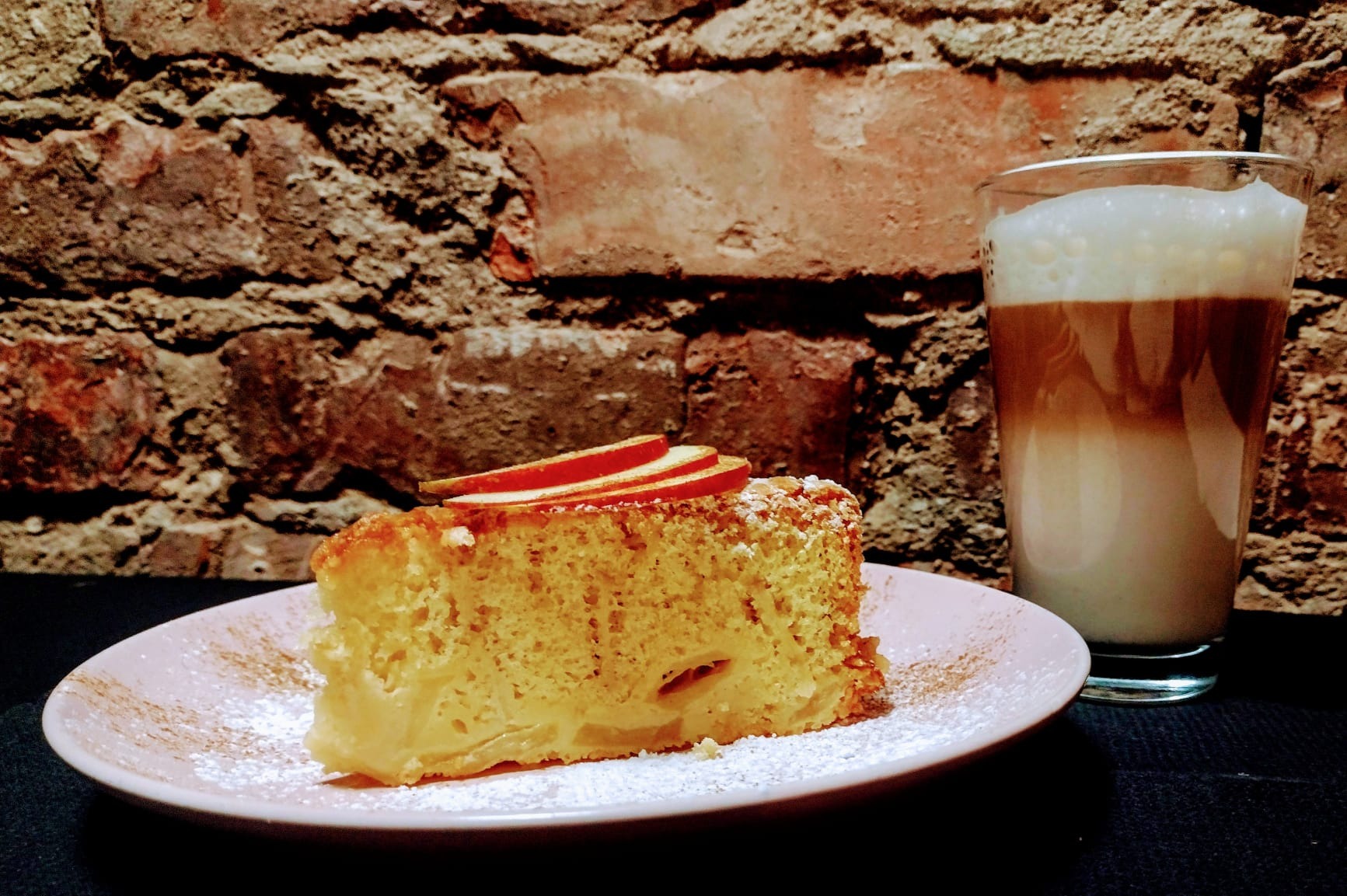 Kawa latte w szklance obok kawałka ciasta jabłkowego z trzema plasterkami jabłka na różowym talerzu, posypane cukrem pudrem, na tle ceglanej ściany.