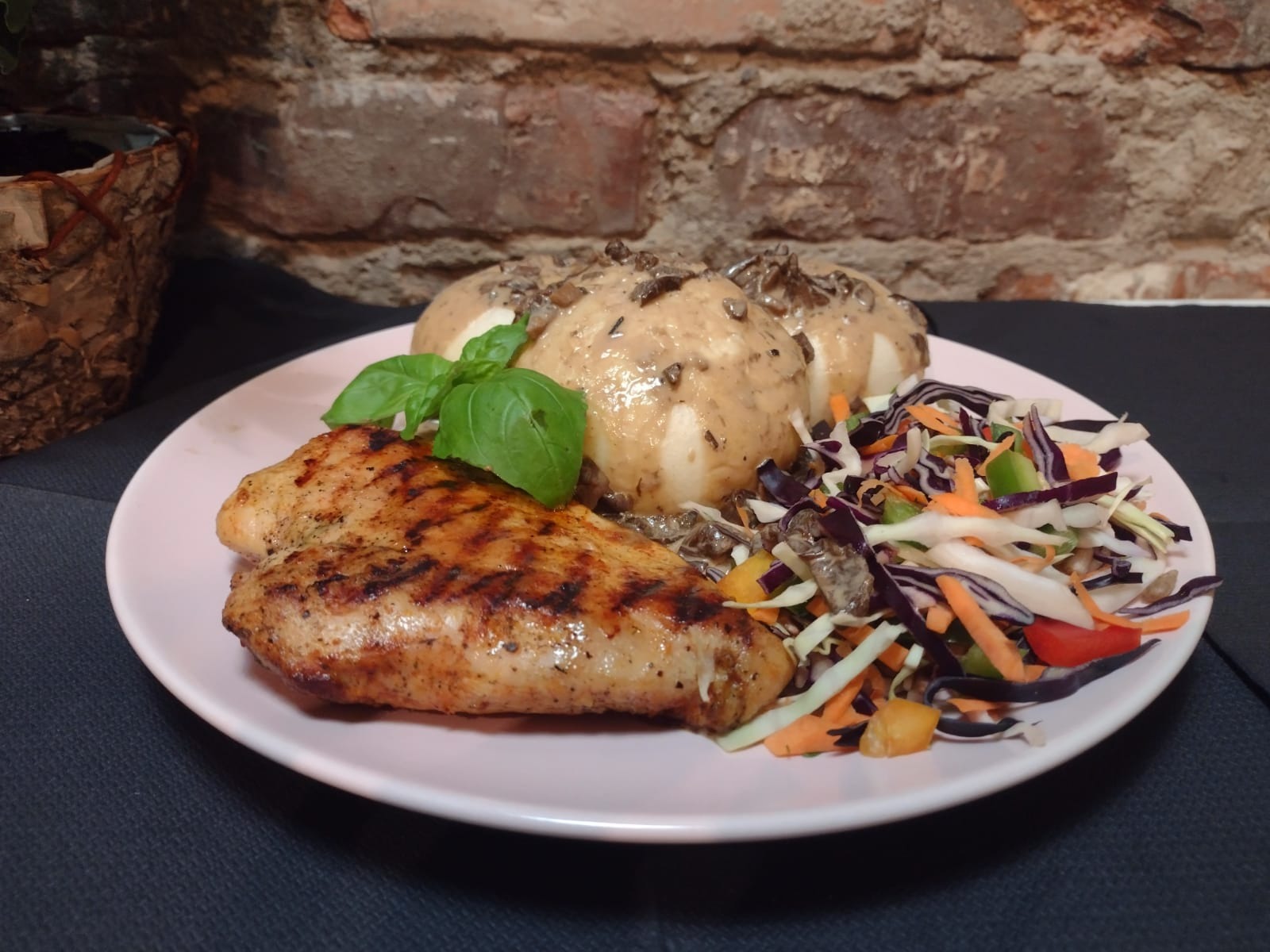 Grillowana pierś kurczaka, trzy ziemniaki w sosie grzybowym i kolorowa surówka na różowym talerzu, bazylia jako dekoracja, na tle ściany z cegły.