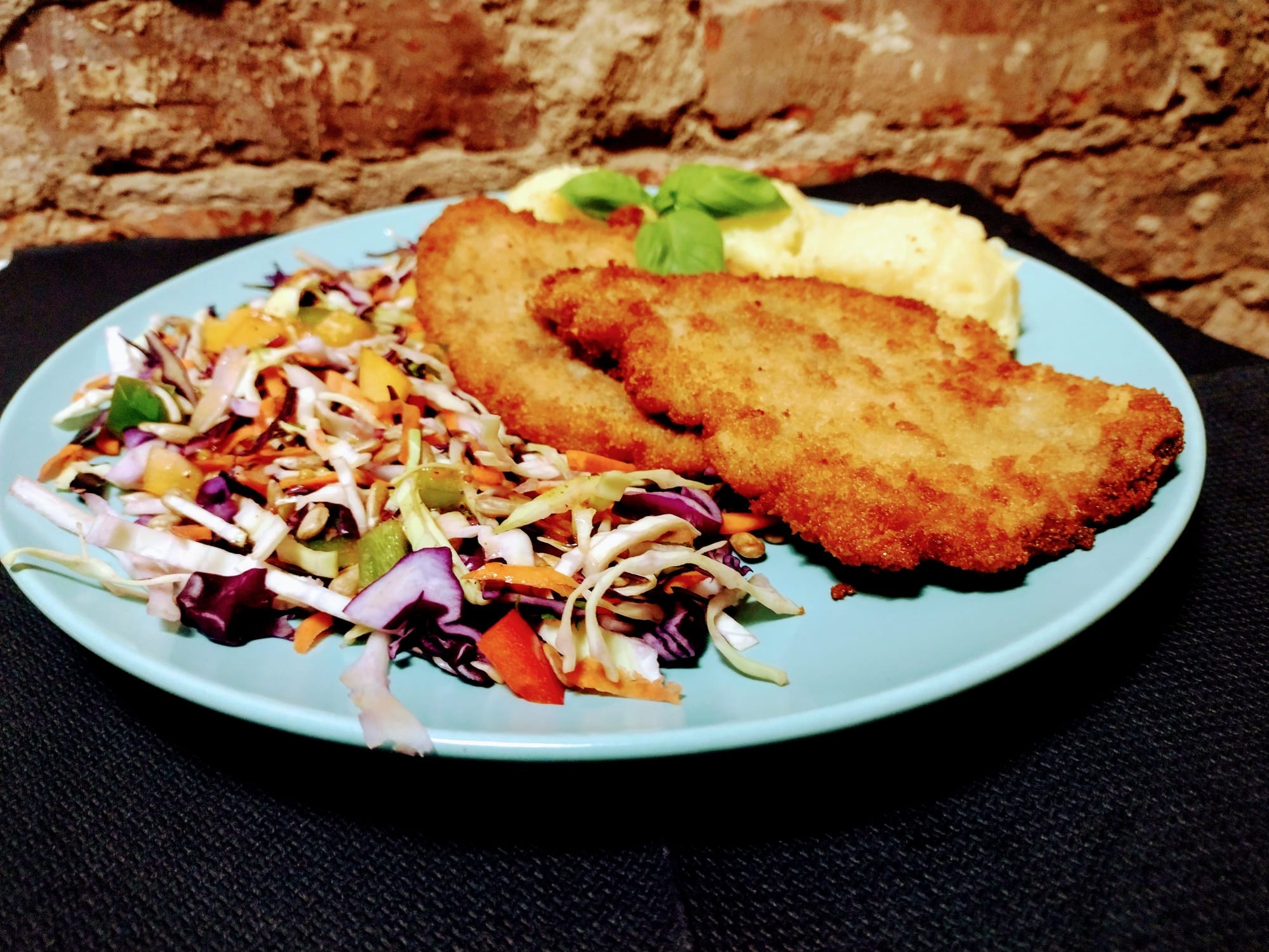 Dwa panierowane kotlety na turkusowym talerzu, podane z puree ziemniaczanym i surówką warzywną; całość udekorowana listkami bazylii, na tle ceglanej ściany.