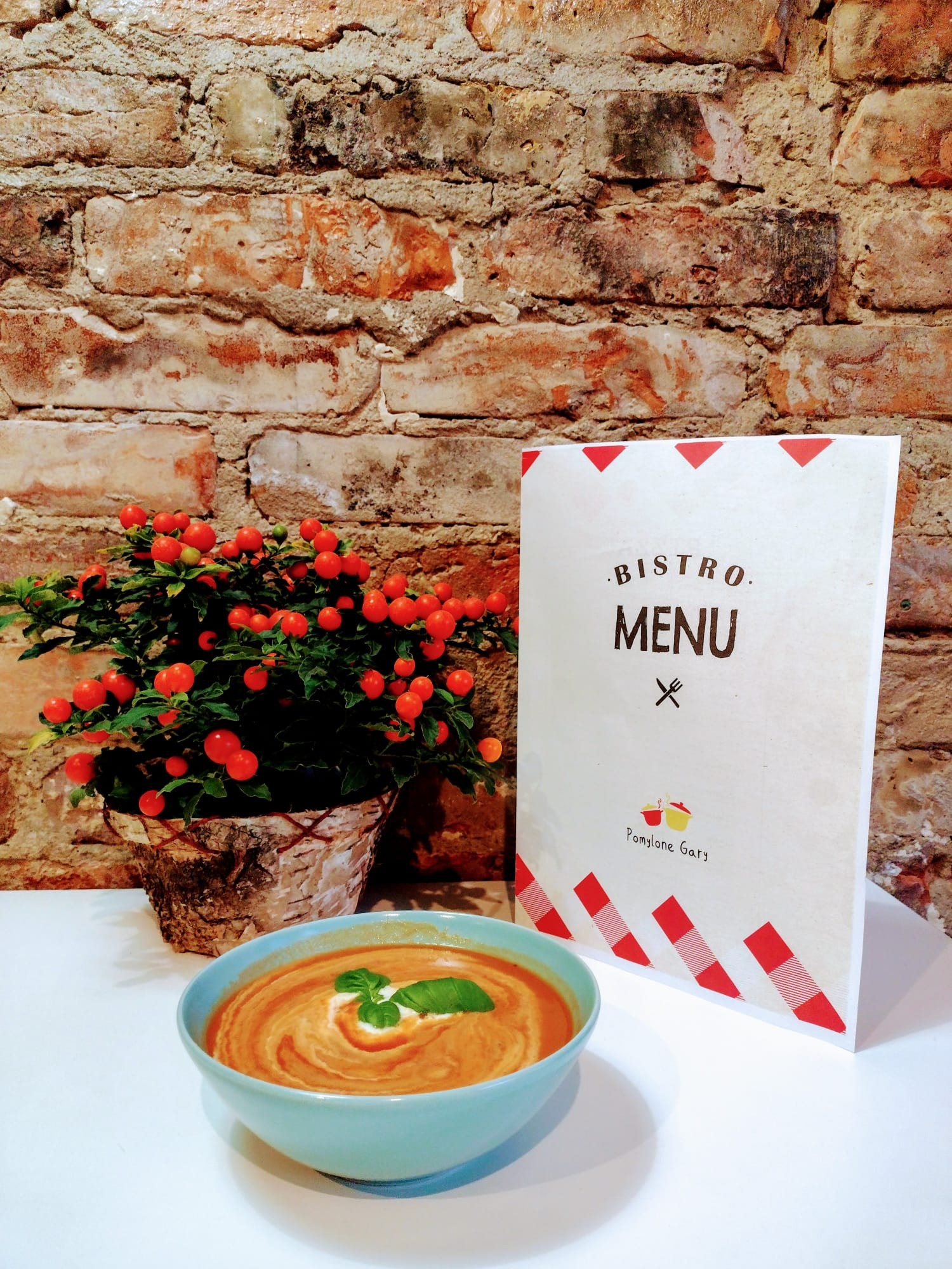 Krem pomidorowy z kleksem śmietany i bazylią w turkusowej miseczce, obok menu bistro i doniczka z kulistymi, czerwonymi owocami na tle ceglanej ściany.