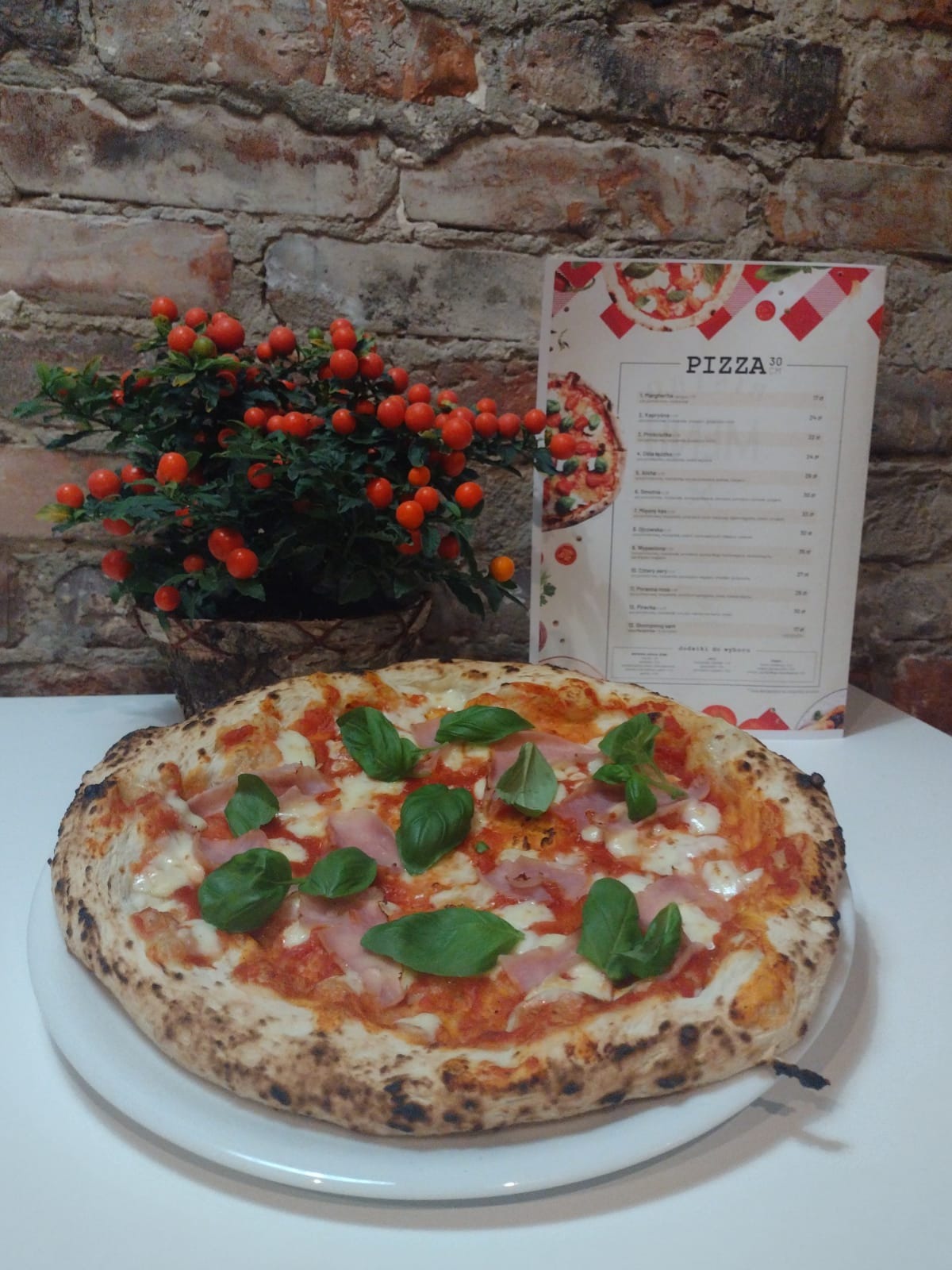 Pizza z szynką i bazylią na białym talerzu, obok roślina z pomarańczowymi owocami i menu, na tle ceglanej ściany.