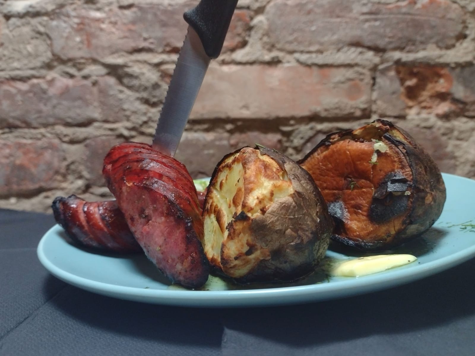 Grillowana kiełbasa z nacięciami, pieczone ziemniaki z ziołami i masłem na turkusowym talerzu, nóż wbity w kiełbasę, na tle ceglanej ściany.
