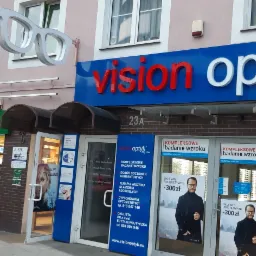 Fasada budynku z szyldem 'Vision Optyk' z dużymi okularami 3D nad wejściem, obok sklep Żabka i drogeria Silcare.