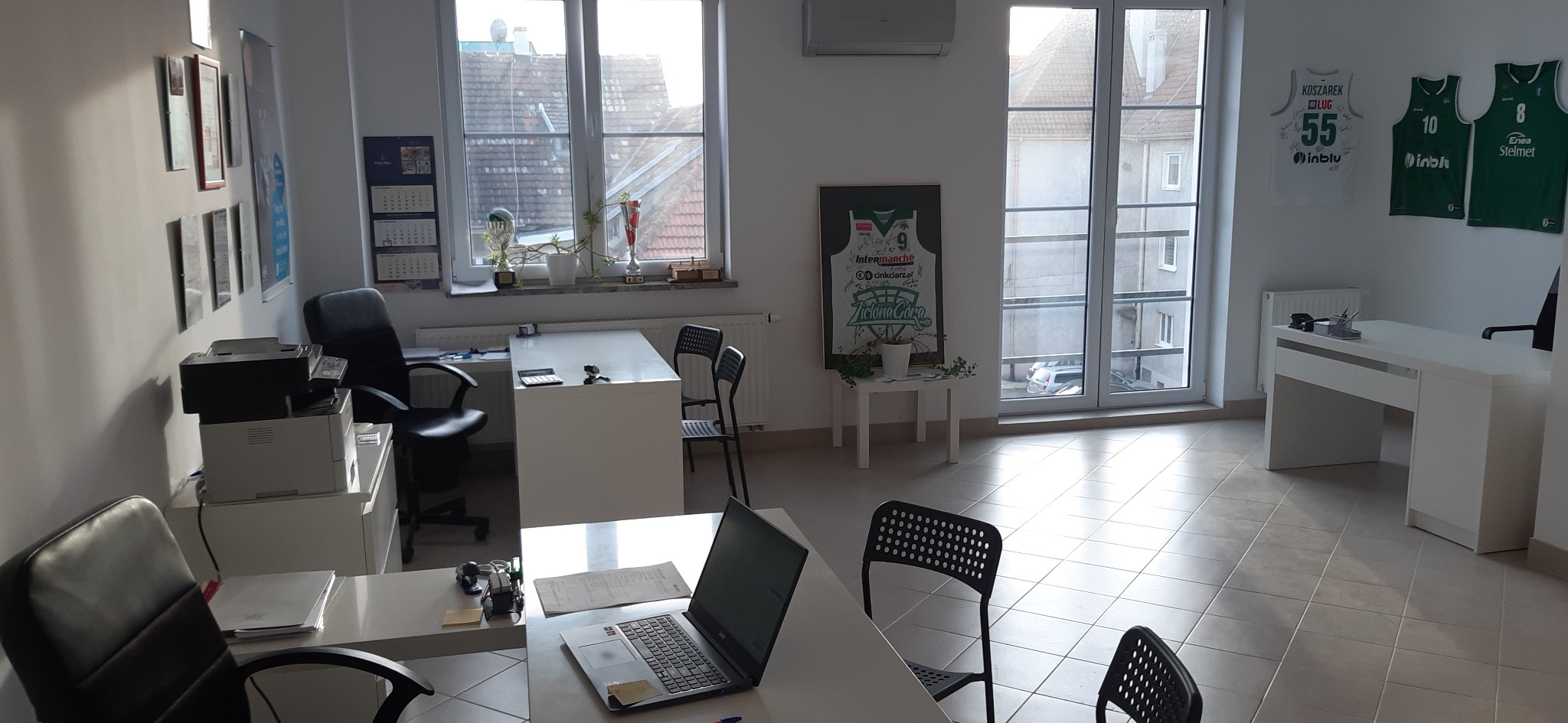 Jasne biuro z biurkami, krzesłami, laptopem, drukarką, kalendarzami, trofeami i oprawionymi koszulkami koszykarskimi na ścianach.