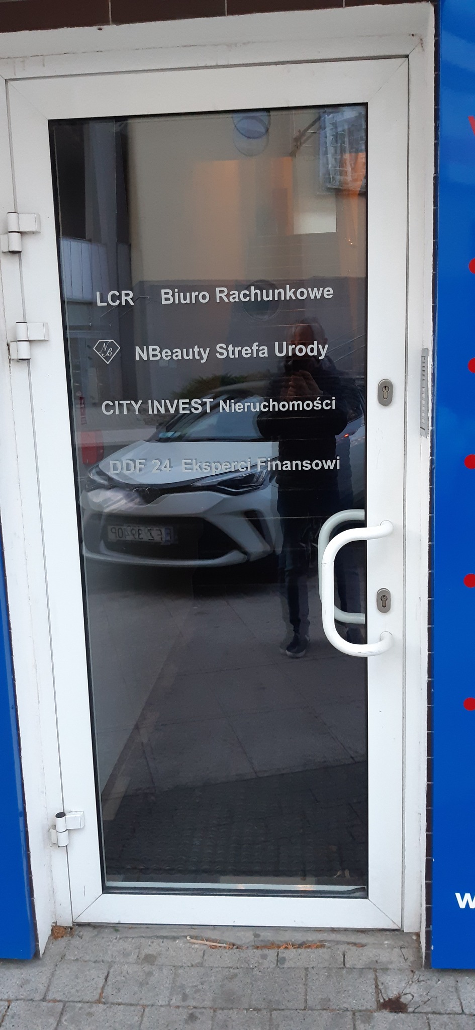 Szklane drzwi wejściowe z napisem 'DDF 24 Eksperci Finansowi', za szybą widoczny fragment białego samochodu.