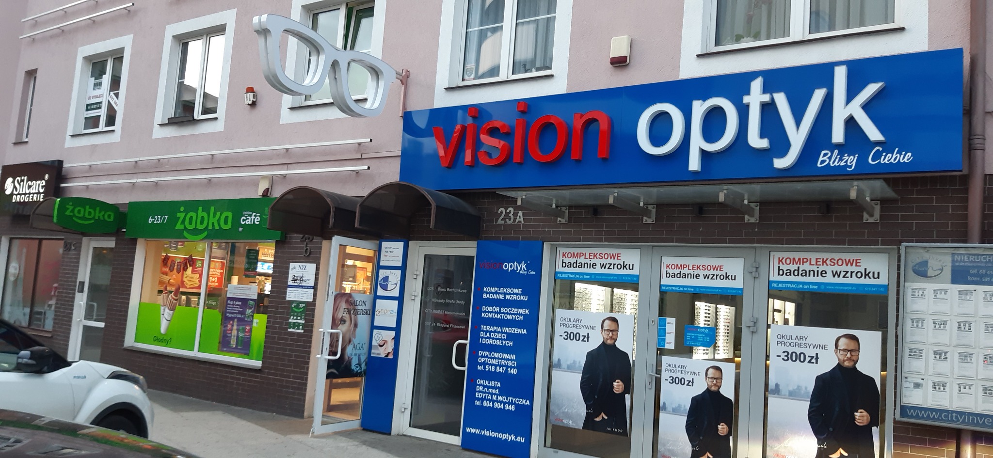 Fasada budynku z szyldem 'Vision Optyk' z dużymi okularami 3D nad wejściem, obok sklep Żabka i drogeria Silcare.