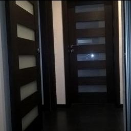 Dwa ciemnobrązowe, nowoczesne drzwi wewnętrzne z przeszkleniami ułożonymi w pionowe pasy. Jedne drzwi otwarte pod kątem, drugie zamknięte. Biała ościeżnica.