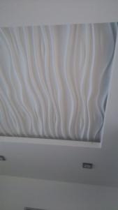 Biały sufit z dekoracyjnymi panelami 3D o falistym wzorze, oświetlenie punktowe w tle.