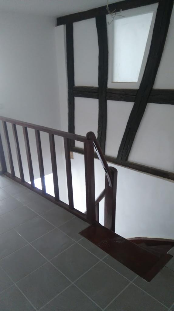 Klatka schodowa z drewnianą balustradą i szarą podłogą z płytek, ściana z elementami drewnianej konstrukcji szkieletowej.