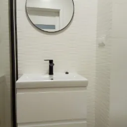 Biała umywalka z czarną baterią, okrągłe lustro w czarnej ramie i białe, teksturowane płytki ścienne tworzą nowoczesną aranżację łazienki.