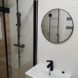 Nowoczesna łazienka z czarną armaturą: kabina prysznicowa typu walk-in z deszczownicą, okrągłe lustro nad białą umywalką z szafką, białe płytki z falistą strukturą.