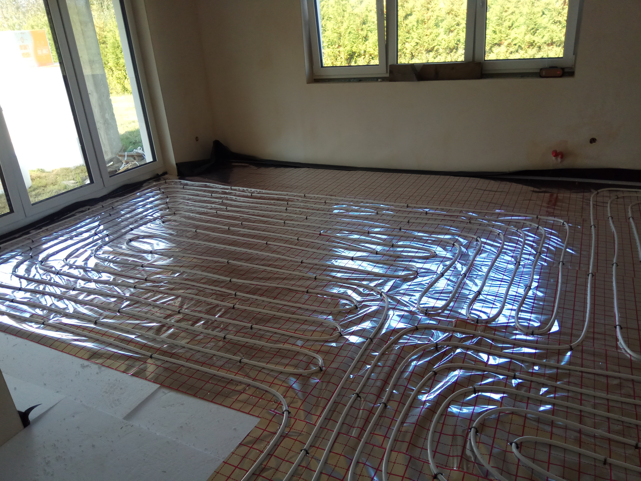 Białe rury ogrzewania podłogowego ułożone na czerwonej siatce w jasnym pomieszczeniu z dużymi oknami, widoczne fragmenty folii i izolacji.