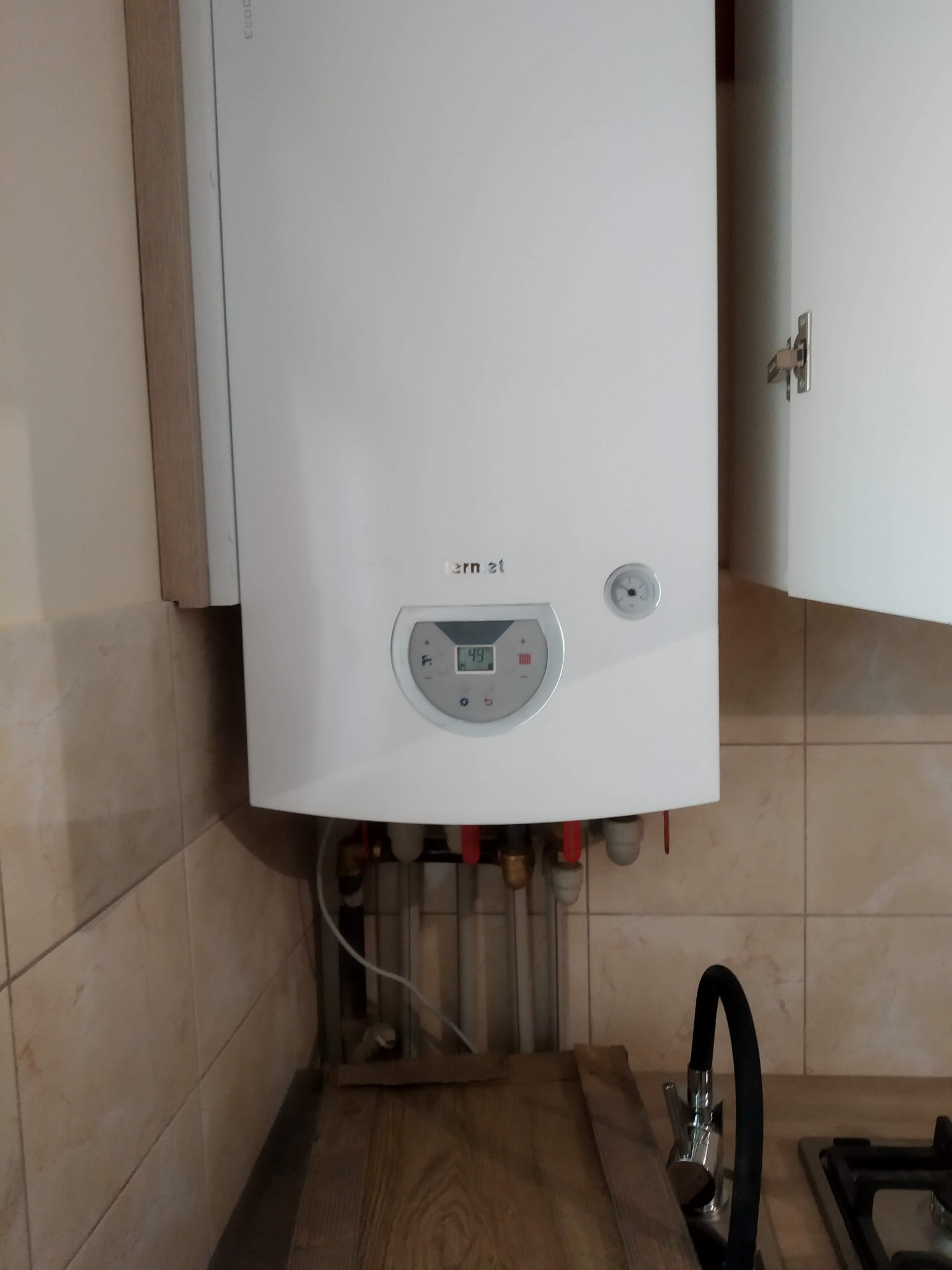 Biały piec gazowy marki Immergas zamontowany w szafce kuchennej, widoczne rury i zawory przyłączeniowe oraz bateria kuchenna z czarną wylewką.