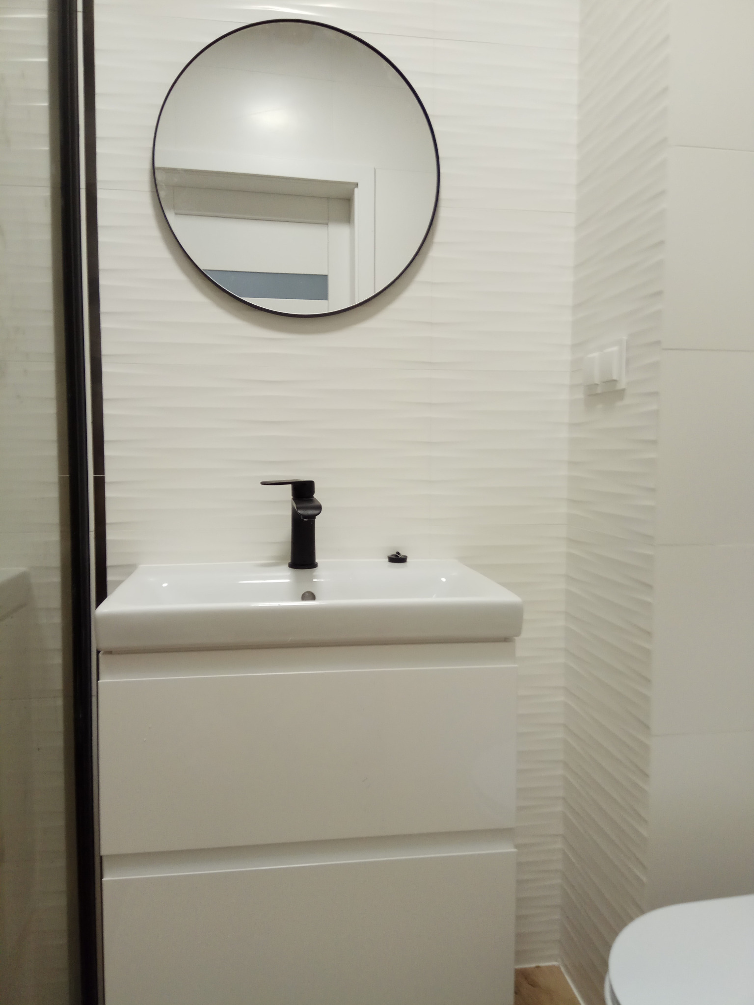 Biała umywalka z czarną baterią, okrągłe lustro w czarnej ramie i białe, teksturowane płytki ścienne tworzą nowoczesną aranżację łazienki.