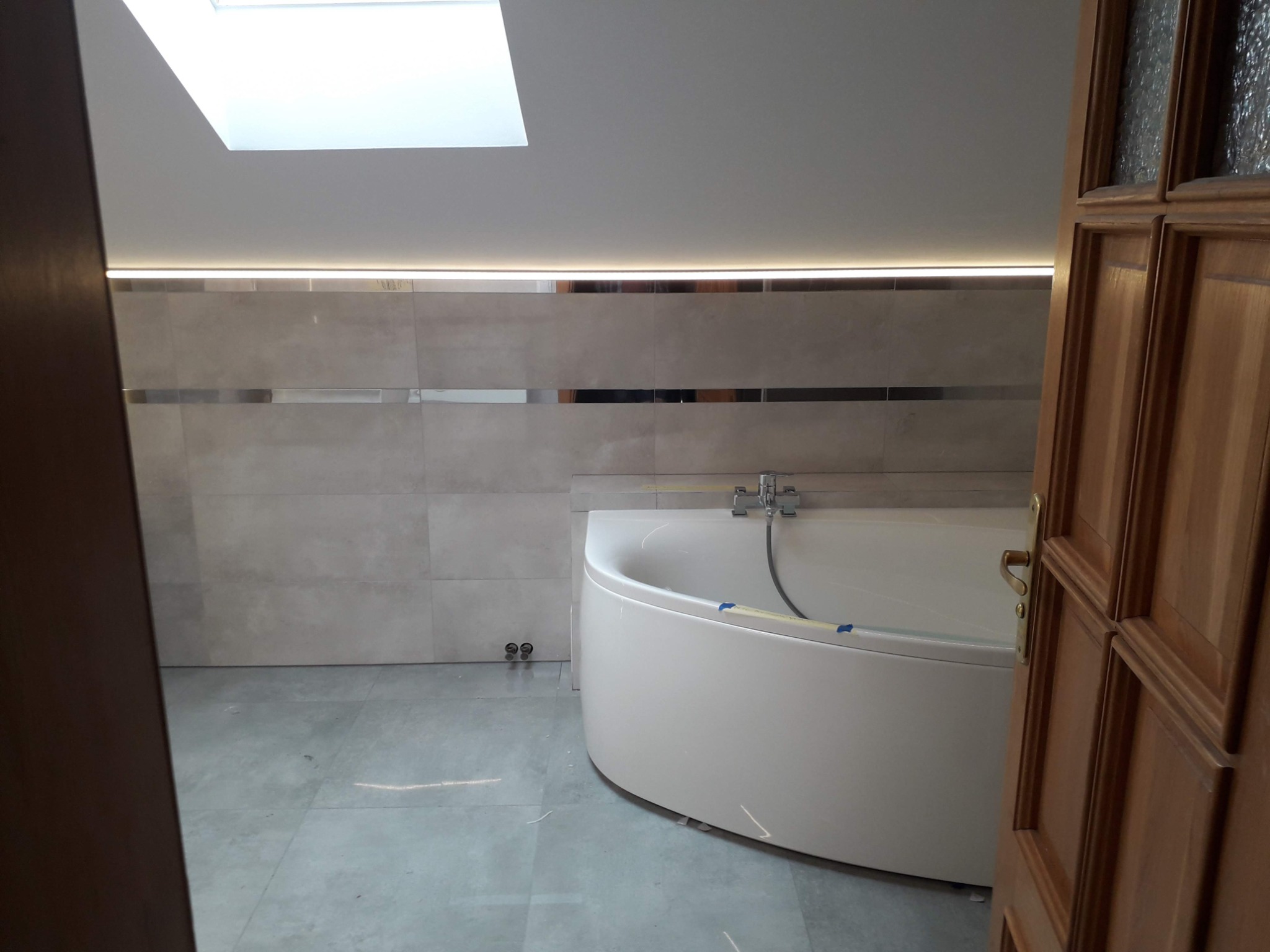 Nowoczesna łazienka z narożną wanną, jasnymi płytkami i oświetleniem LED pod sufitem, widok zza drewnianych drzwi.