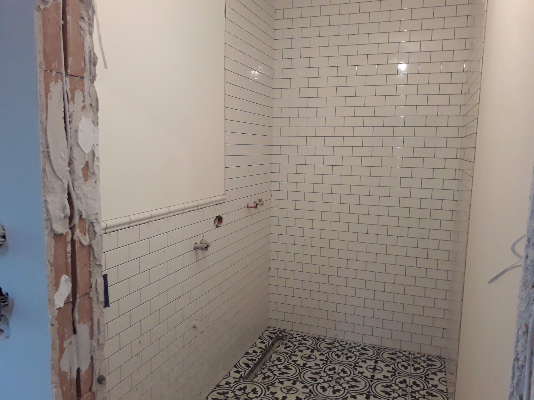 Świeżo wykafelkowana łazienka z białymi, prostokątnymi płytkami na ścianach i wzorzystą, czarno-białą podłogą; widoczne wyprowadzenia instalacji wodnej i surowy fragment ściany w trakcie prac...