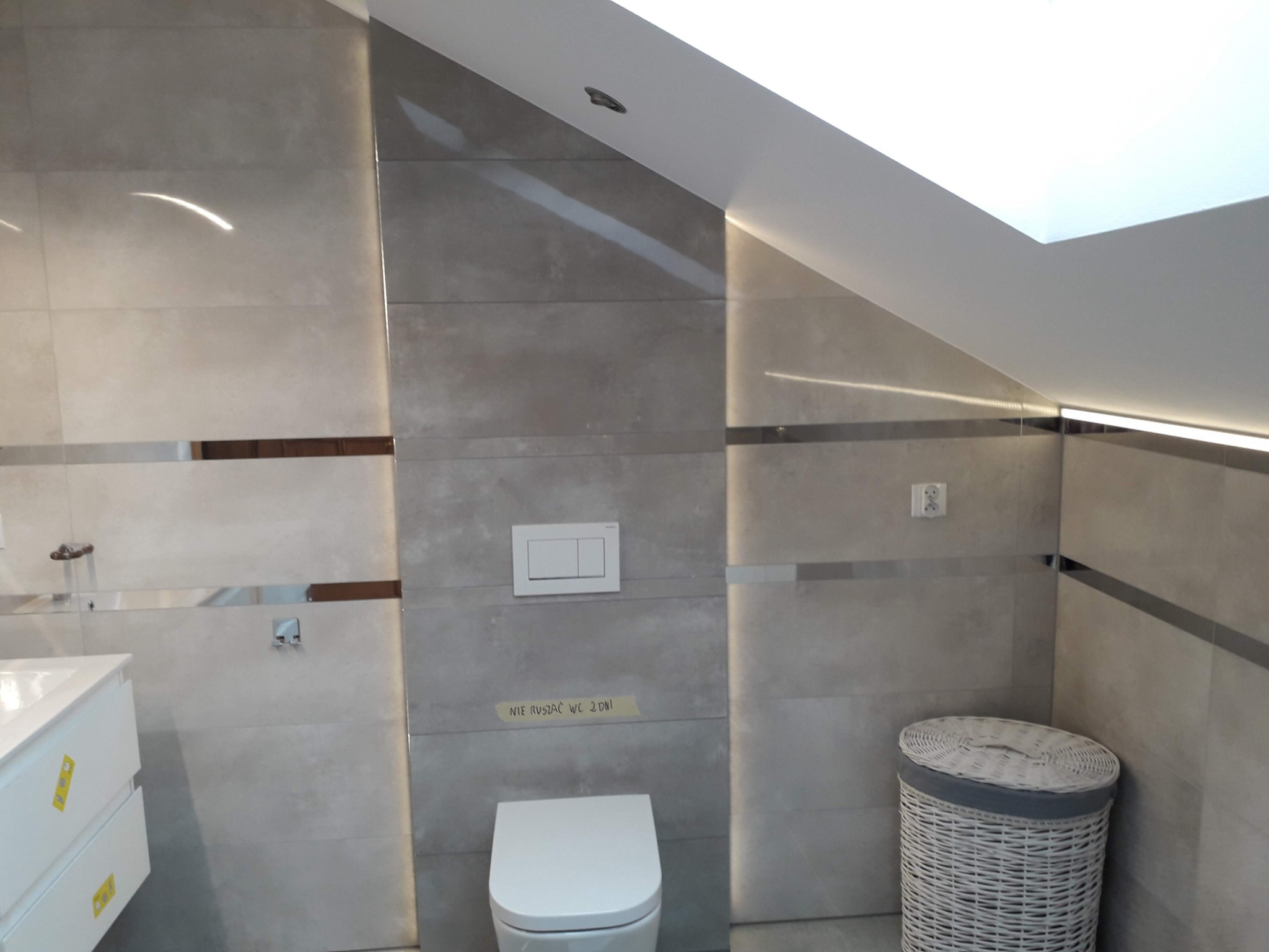 Jasna łazienka na poddaszu ze skośnym sufitem, wykończona płytkami imitującymi beton z dekoracyjnymi listwami, biała umywalka z szafką, toaleta i kosz na pranie.