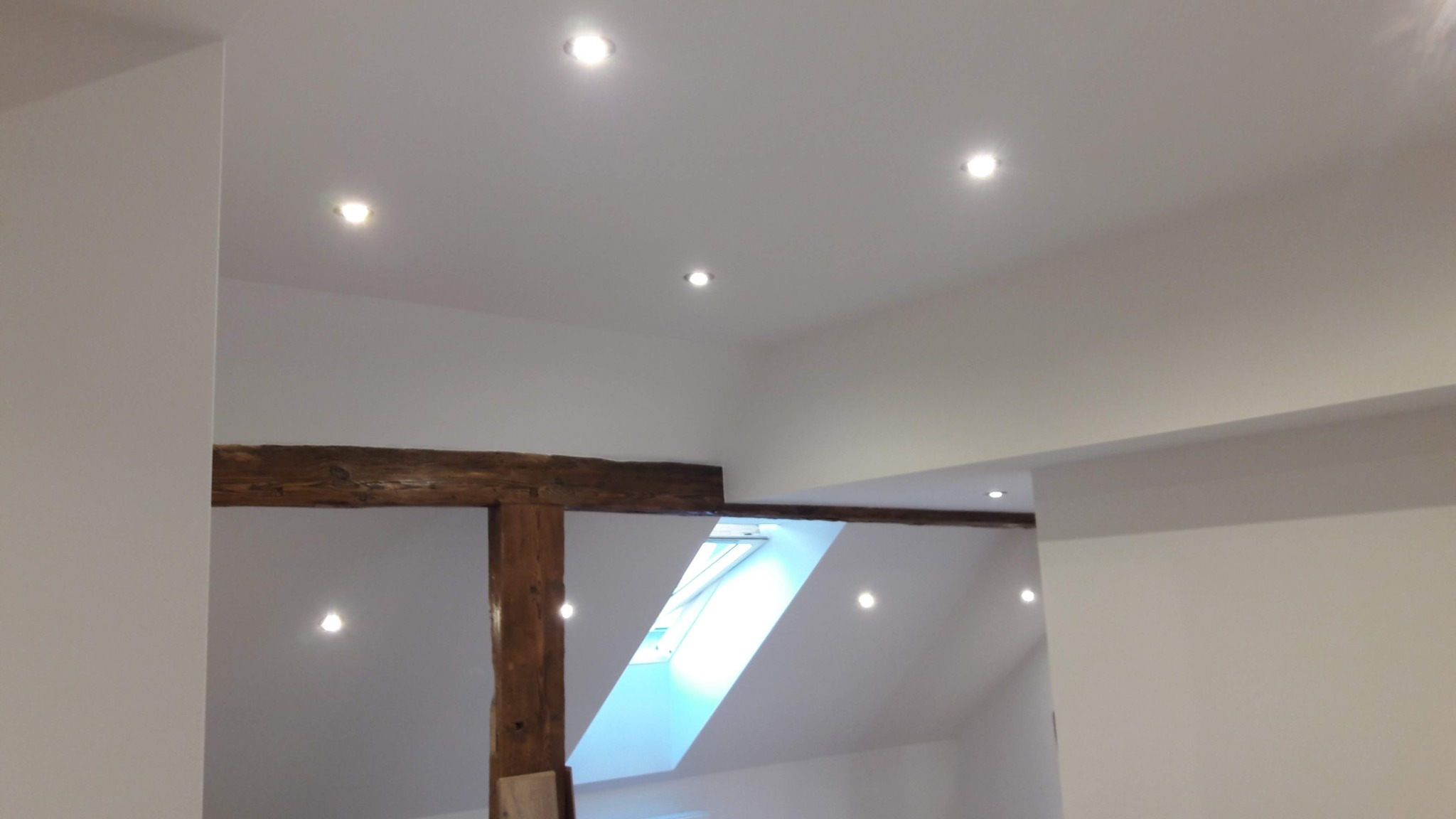 Wnętrze poddasza po remoncie z drewnianą belką, oświetleniem punktowym i oknem dachowym, wykończone na biało.
