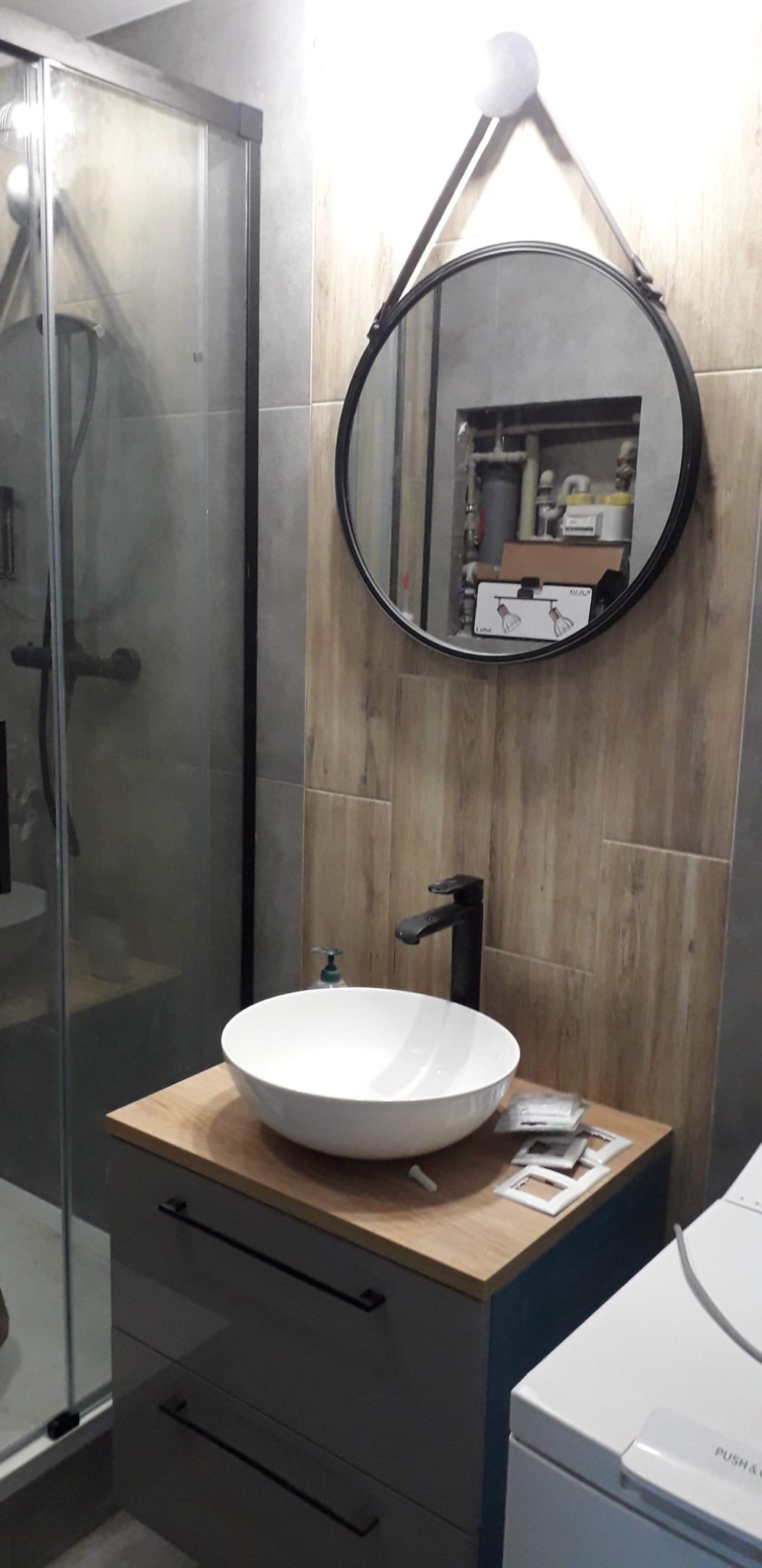 Nowoczesna łazienka z okrągłym lustrem na pasku, umywalką nablatową i szarą szafką z ciemnymi uchwytami, obok toaleta i kabina prysznicowa z czarnymi profilami.