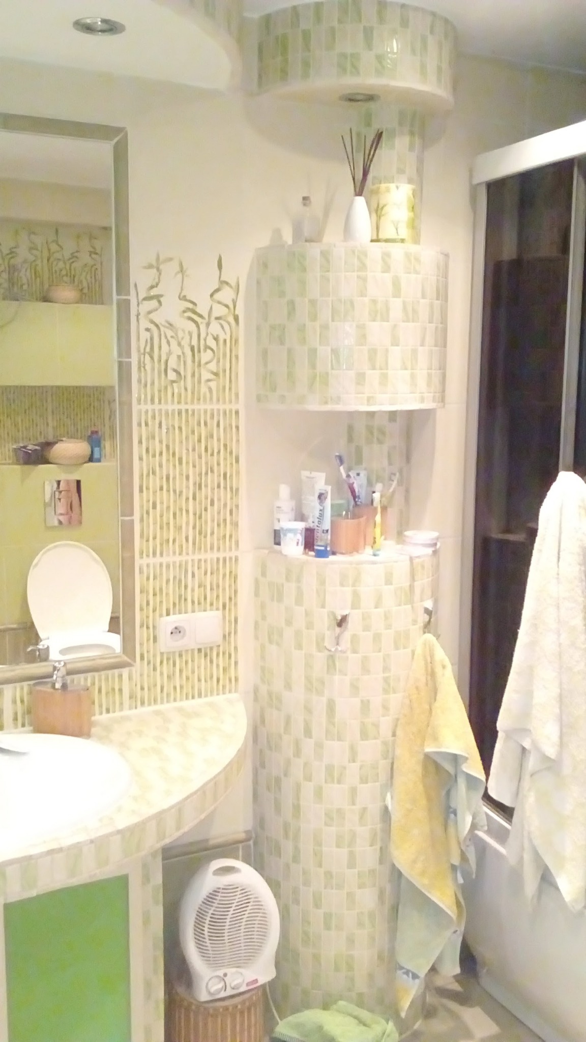 Ukończona łazienka z zielonymi akcentami: mozaika na ścianach, toaleta z podświetleniem, umywalka z blatem, dekoracje roślinne i grzejnik na wiklinowym stołku.