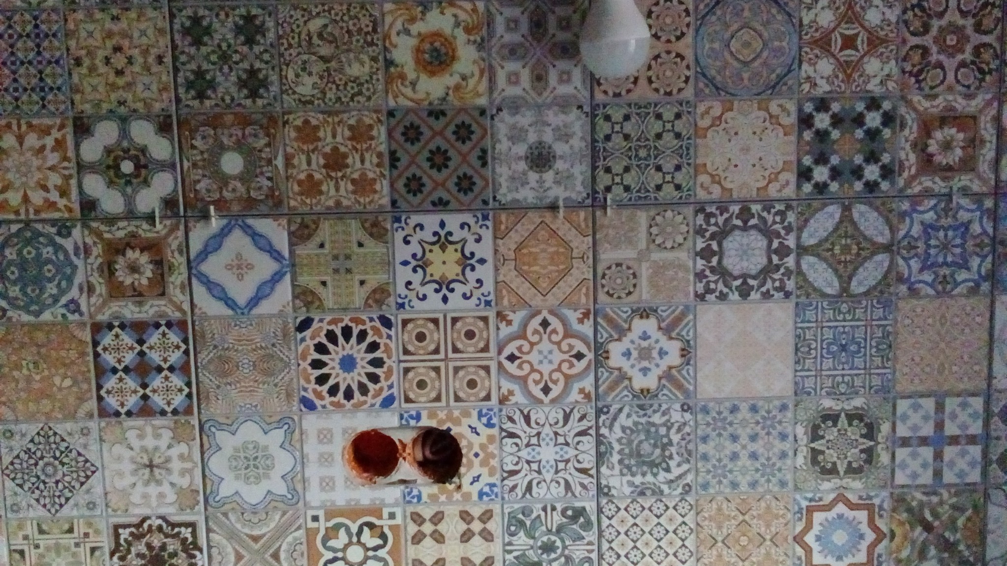 Ściana wyłożona różnorodnymi, wzorzystymi płytkami ceramicznymi w stylu patchwork, z widocznym otworem na instalację elektryczną i żarówką w górnym rogu.