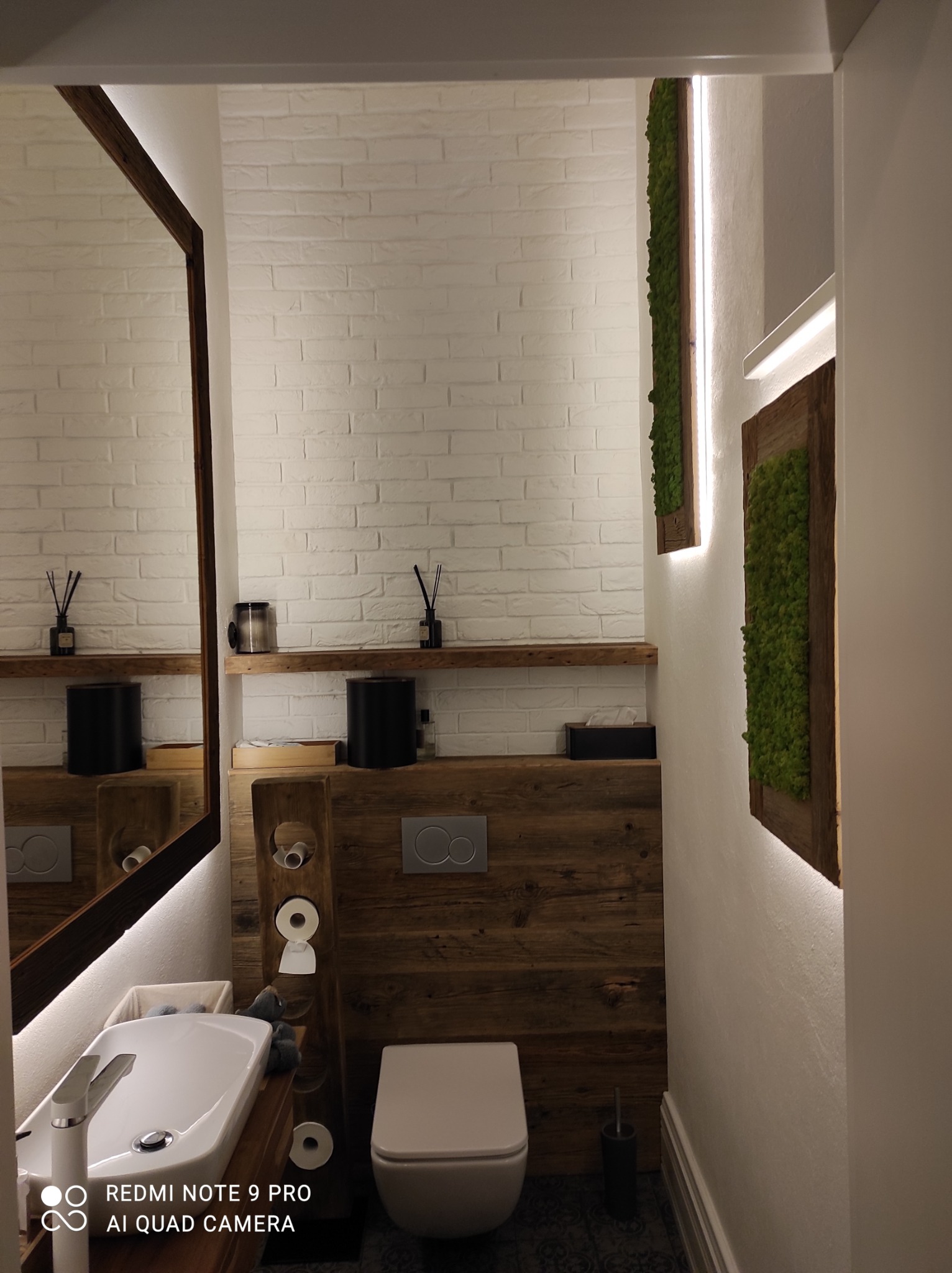 Nowoczesna łazienka z białymi cegiełkami, drewnianymi akcentami i panelami z mchu podświetlonymi LED, widok na umywalkę, toaletę i lustro.