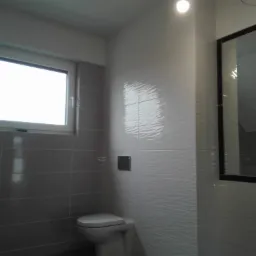 Wykończona łazienka z szarymi i białymi płytkami, wisząca toaleta, okno i duże lustro z czarną ramą, widoczne odbicie ręki na lustrze.