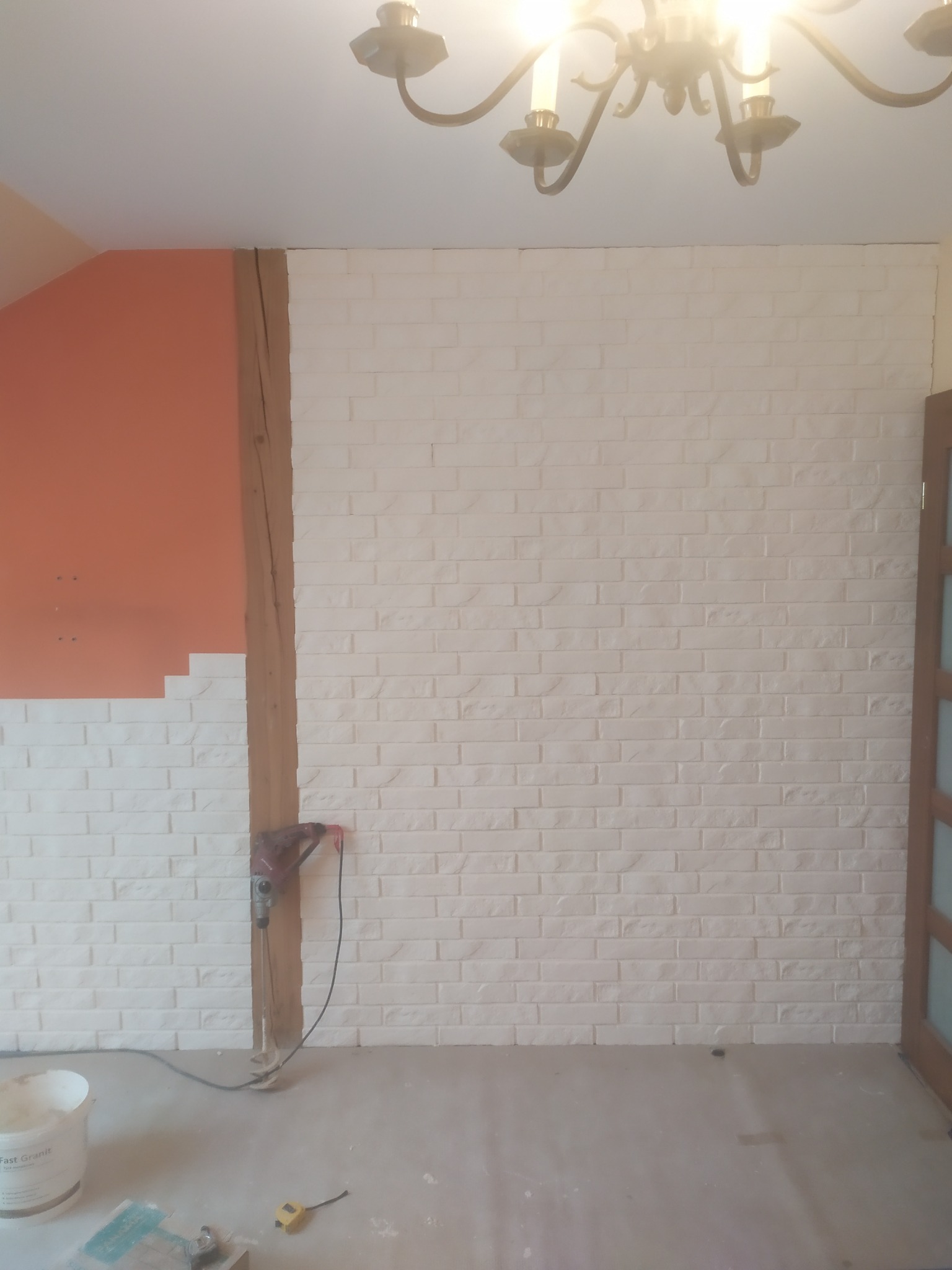 Ściana z białej cegły dekoracyjnej w trakcie montażu w Górze, widoczny fragment pomalowanej na pomarańczowo ściany i narzędzia.