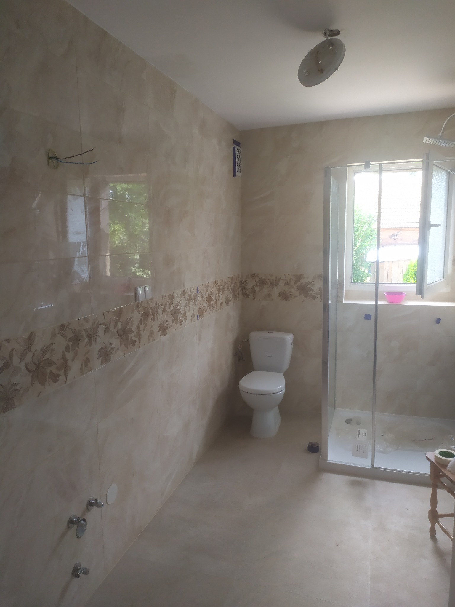 Ukończona łazienka z beżowymi płytkami z kwiatowym motywem, kabiną prysznicową z przezroczystego szkła, białą toaletą i otwartym oknem z widokiem na zieleń.